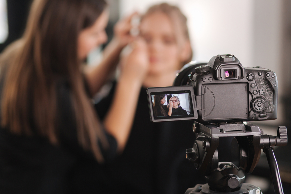 Video camera in foreground with makeup artist and seated client in background